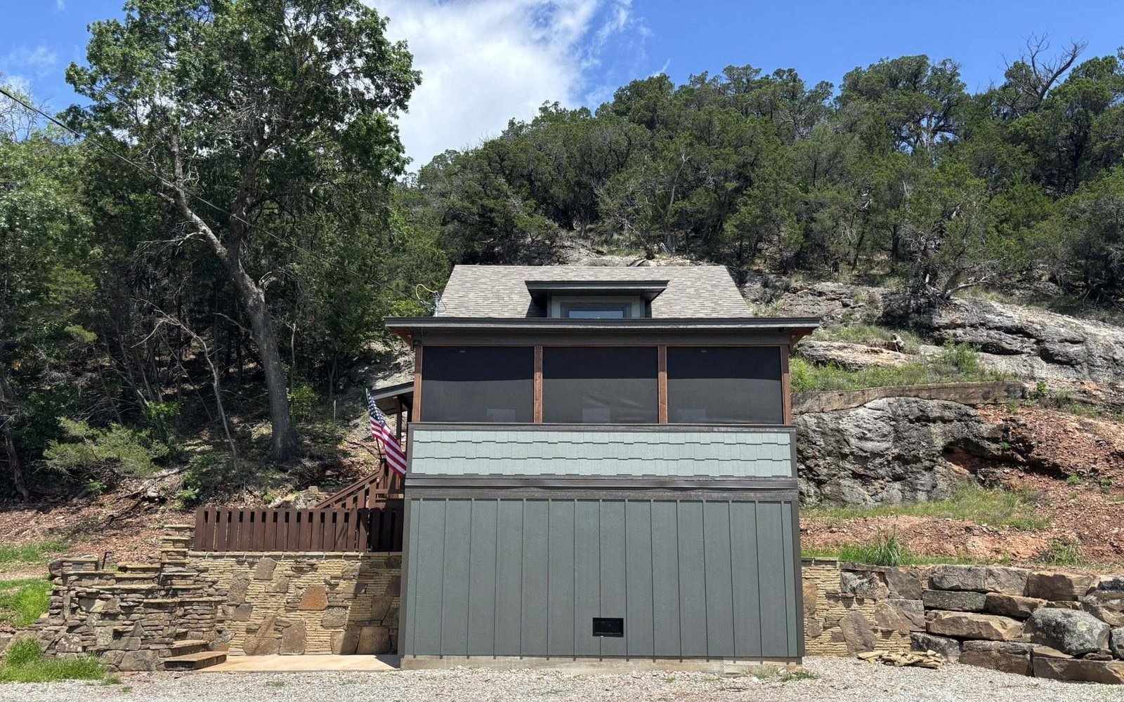 The cabin seen from the parking area — green siding, screened upper porch, stone retaining walls into the hillside