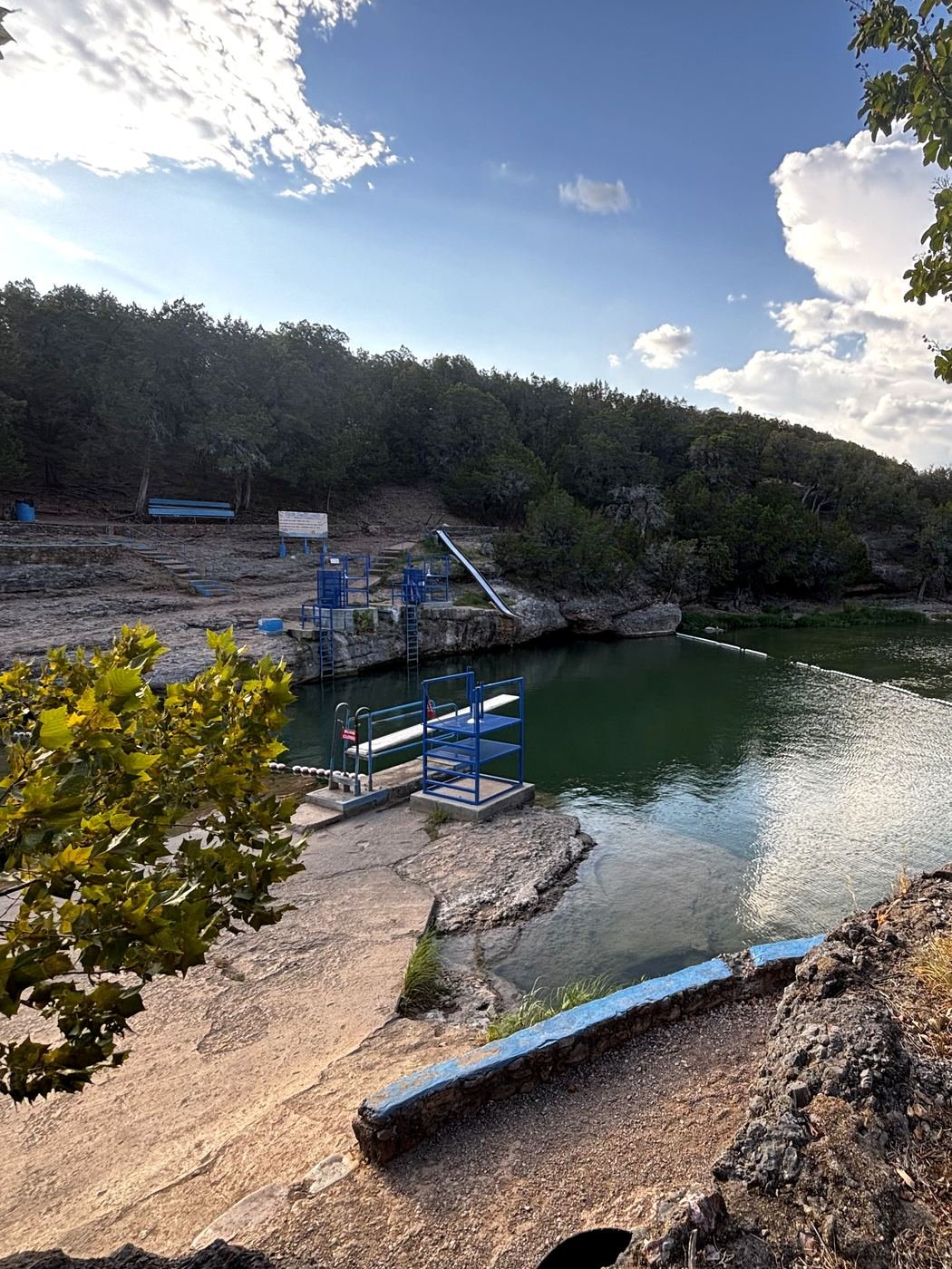 The Blue Hole at Turner Falls Park: the slide and diving platforms above a green pool, with a shallow rock shelf and wooded bluffs behind